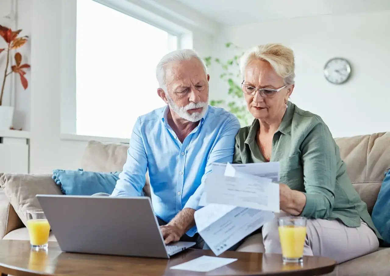 Couple âgé examinant des documents, peut-être pour une exonération taxe foncière 2026. Ils sont sur un canapé avec un ordinateur portable et des boissons.