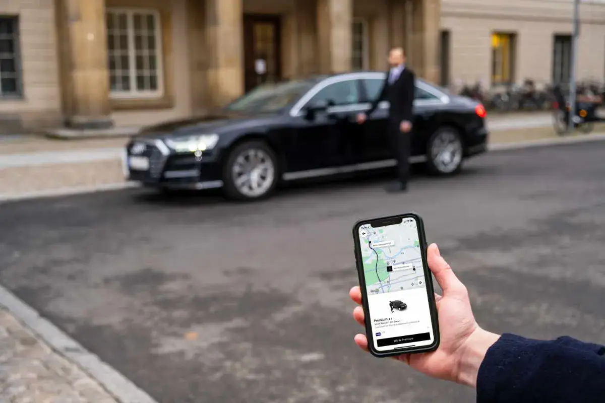 Hand h&auml;lt Smartphone mit Uber-App, die Uber Premium Cars anzeigt. Ein Mann in Anzug steht neben einem schwarzen Luxusauto.