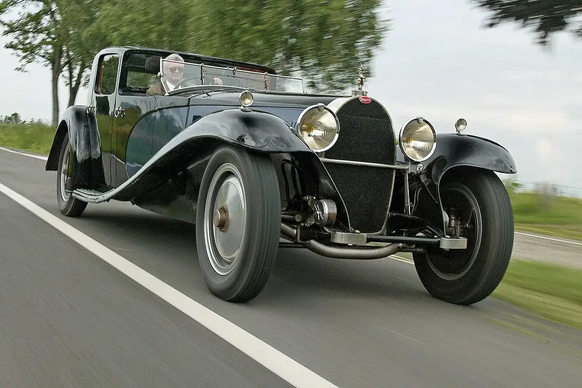 Ein Mann f&auml;hrt einen gl&auml;nzenden schwarzen Bugatti Royale auf einer Landstra&szlig;e. Der Bugatti Royale Preis ist astronomisch, ein Traum f&uuml;r jeden Sammler.