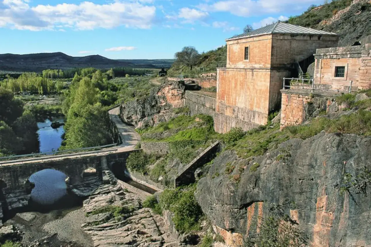Antiguo pontón de la oliva junto a un río y un puente de piedra, con edificios históricos en la ladera rocosa.
