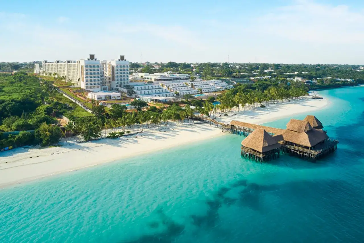 Rajski widok na Riu Jambo Zanzibar: biała plaża, turkusowa woda i hotel z pomostem.
