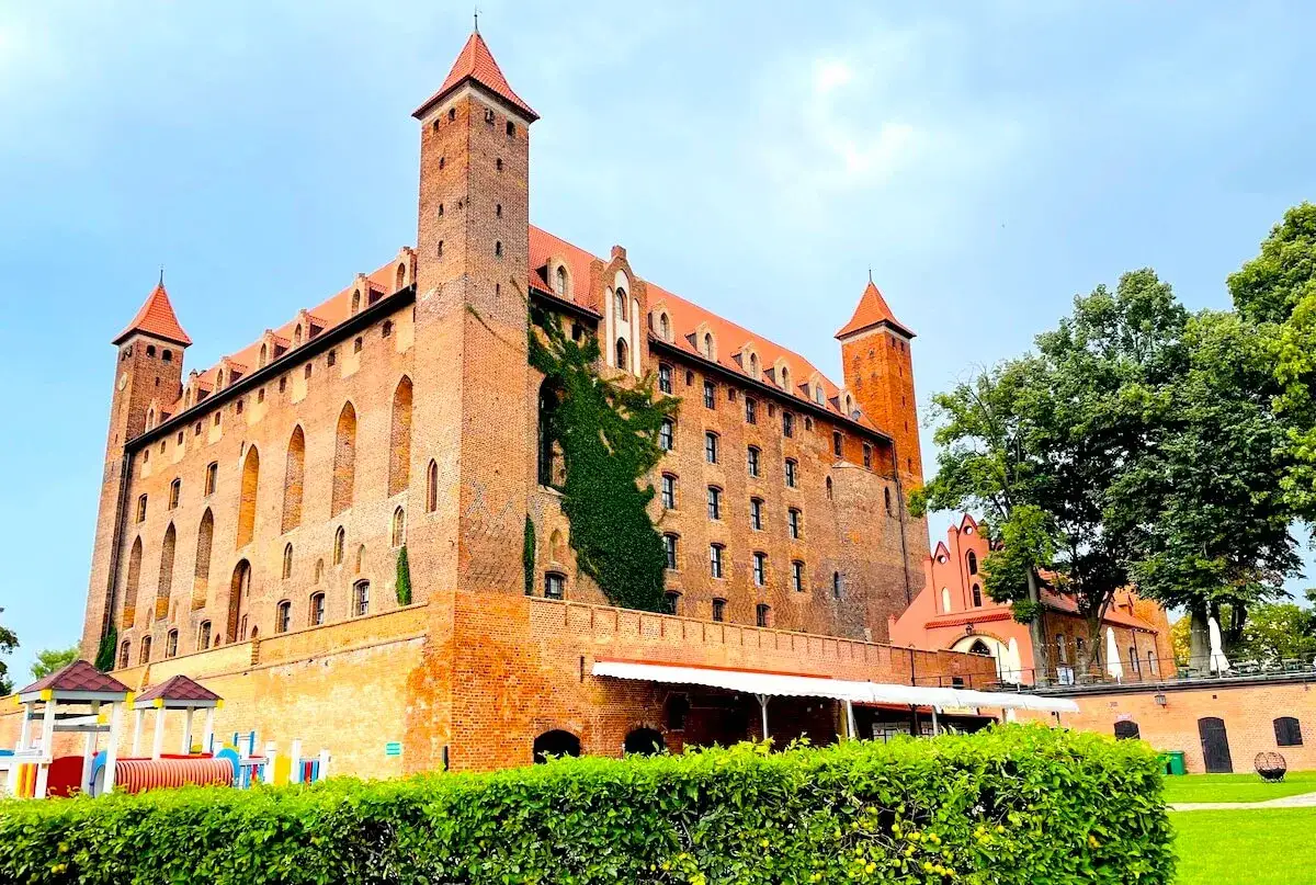 Imponujące zamki pomorskie z cegły, z wieżami i zielonym bluszczem, na tle błękitnego nieba. Obok plac zabaw i zadbane ogrody.