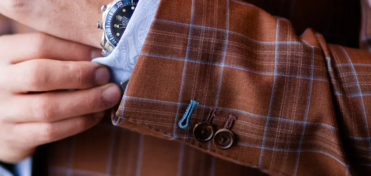 A man adjusts his watch on his wrist, revealing the detailed pick stitching on the cuff of his brown plaid suit jacket.