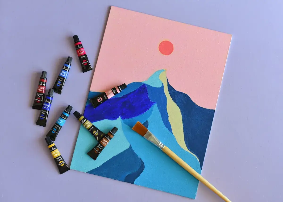 Un tableau abstrait de montagnes roses et bleues, avec des tubes de peinture acrylique et un pinceau. Une image à peindre facile pour débuter.