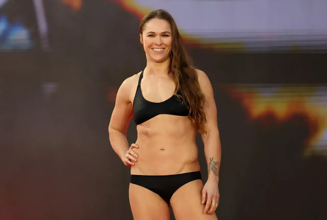 Ronda Rousey smiles, wearing a black bikini. She's a champion who has fought in various weight brackets.