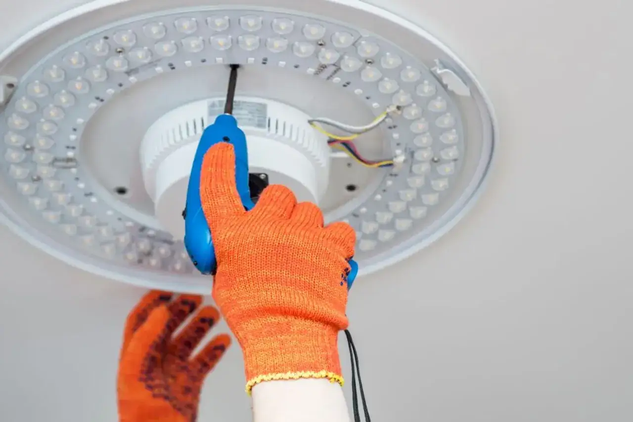 Ręka w pomarańczowej rękawicy przykręca śrubokrętem nowoczesny plafon sufitowy LED. Poradnik: jak zamontować plafon sufitowy.