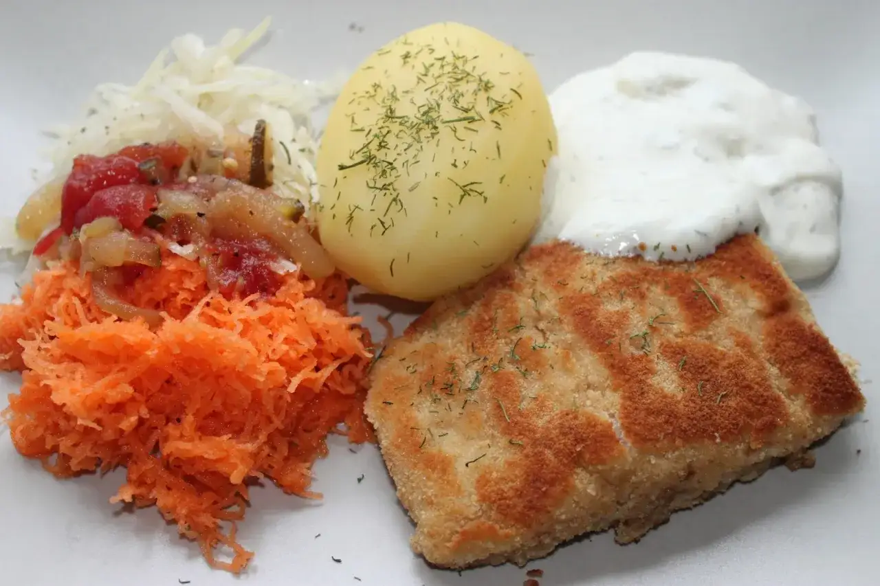 En gyllene panerad fisk med dill, en kokt potatis, vit sås och en sallad på riven morot och surkål.