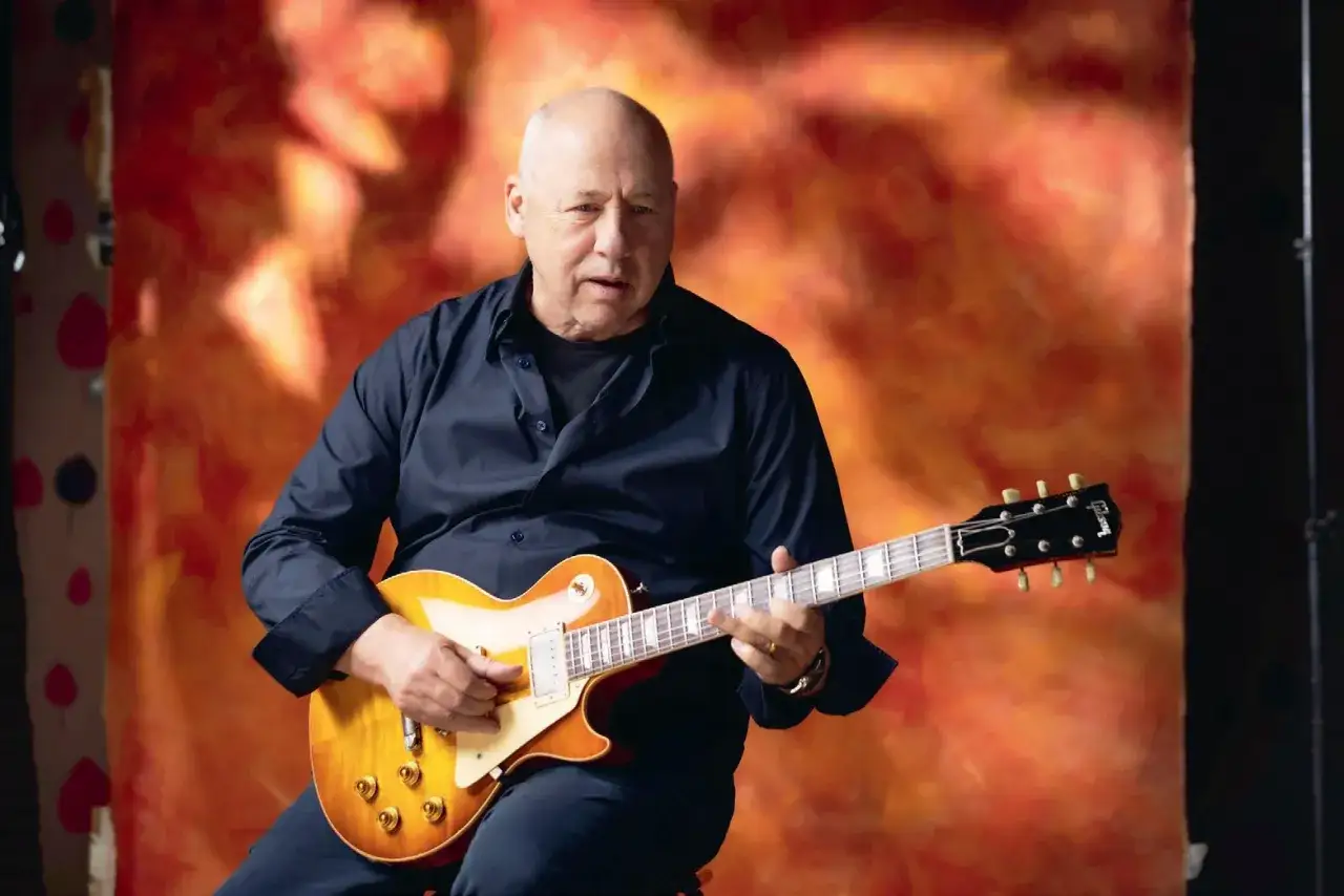 Mark Knopfler, legenda gitary, przygotowuje się do kolejnych koncertów. Gra na swoim ukochanym Gibsonie na tle ognistego tła.