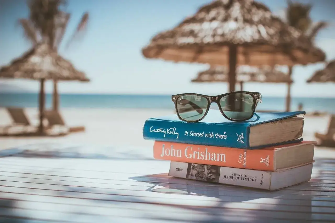 Occhiali da sole su una pila di libri da leggere in estate, con spiaggia e mare sullo sfondo.