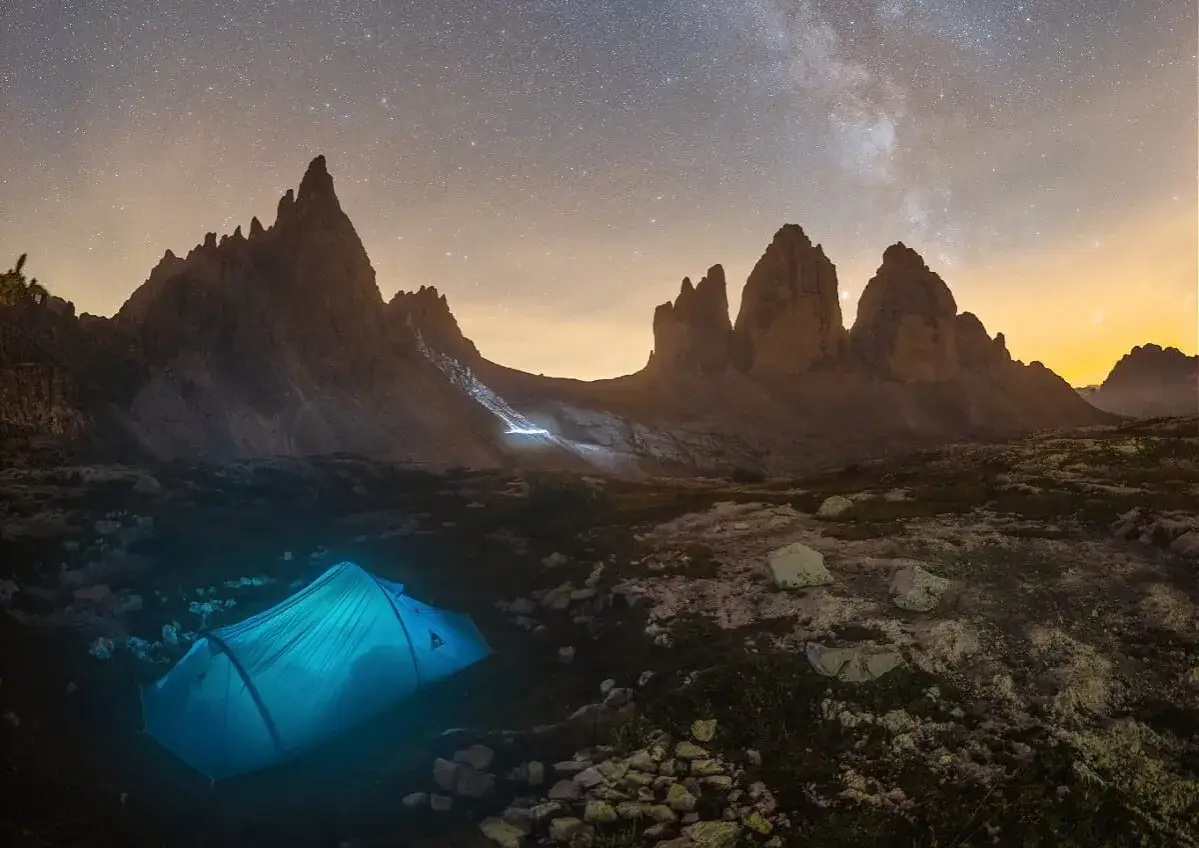 Nocny camping rocchetta pod rozgwieżdżonym niebem. Błękitny namiot świeci w mroku, a za nim wznoszą się skaliste szczyty.