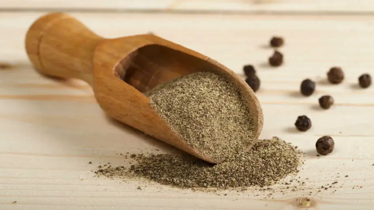 Cuchara de madera con pimienta negra molida y granos enteros esparcidos. Descubre las **pimienta negra propiedades** y su uso en la cocina.