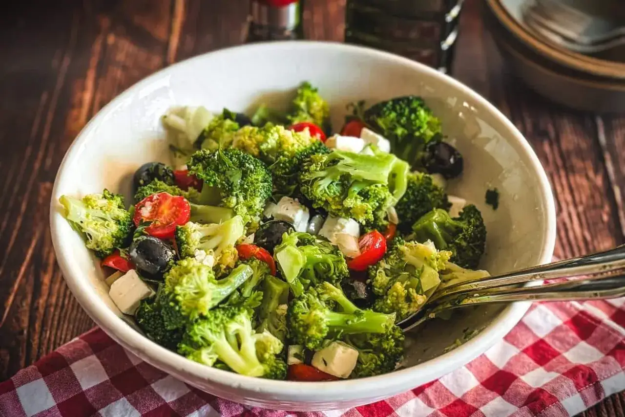 Frischer brokkoli salat mit Kirschtomaten, schwarzen Oliven und Feta-Würfeln in einer weißen Schüssel, serviert auf kariertem Stoff.
