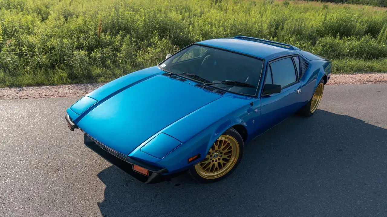 Błękitny De Tomaso Pantera z lat 70. na złotych felgach, zaparkowany na poboczu drogi, z zielonym polem w tle.