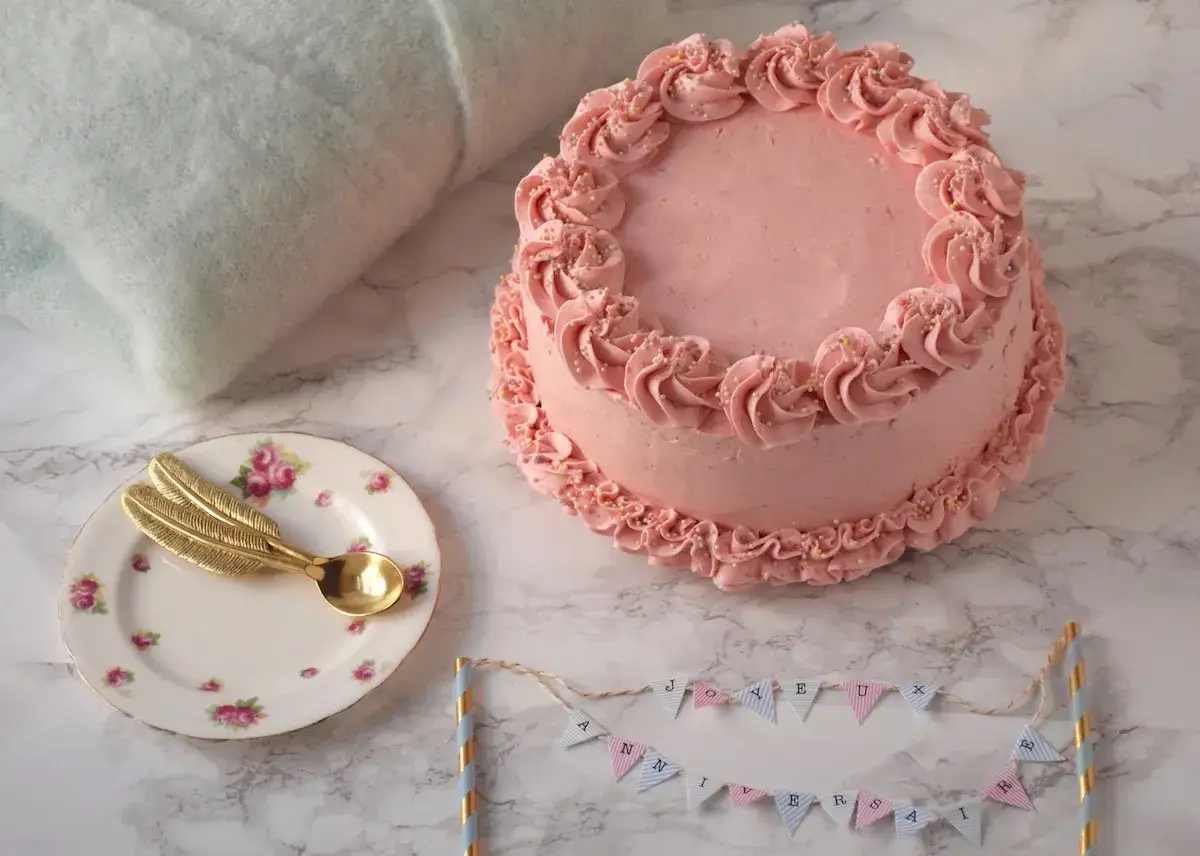 Gâteau rose pastel, décoré de volutes et de perles. Une petite assiette avec une cuillère dorée en forme de plume et une guirlande 