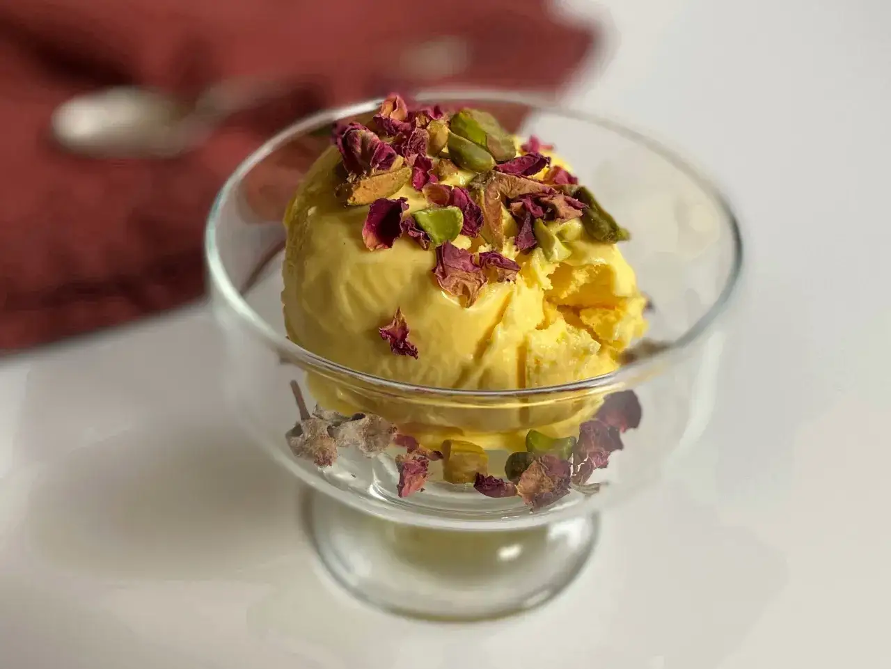 Ein Löffel cremiges, gelbes persisches Eis, garniert mit Pistazien und Rosenblättern, in einem Glas serviert.
