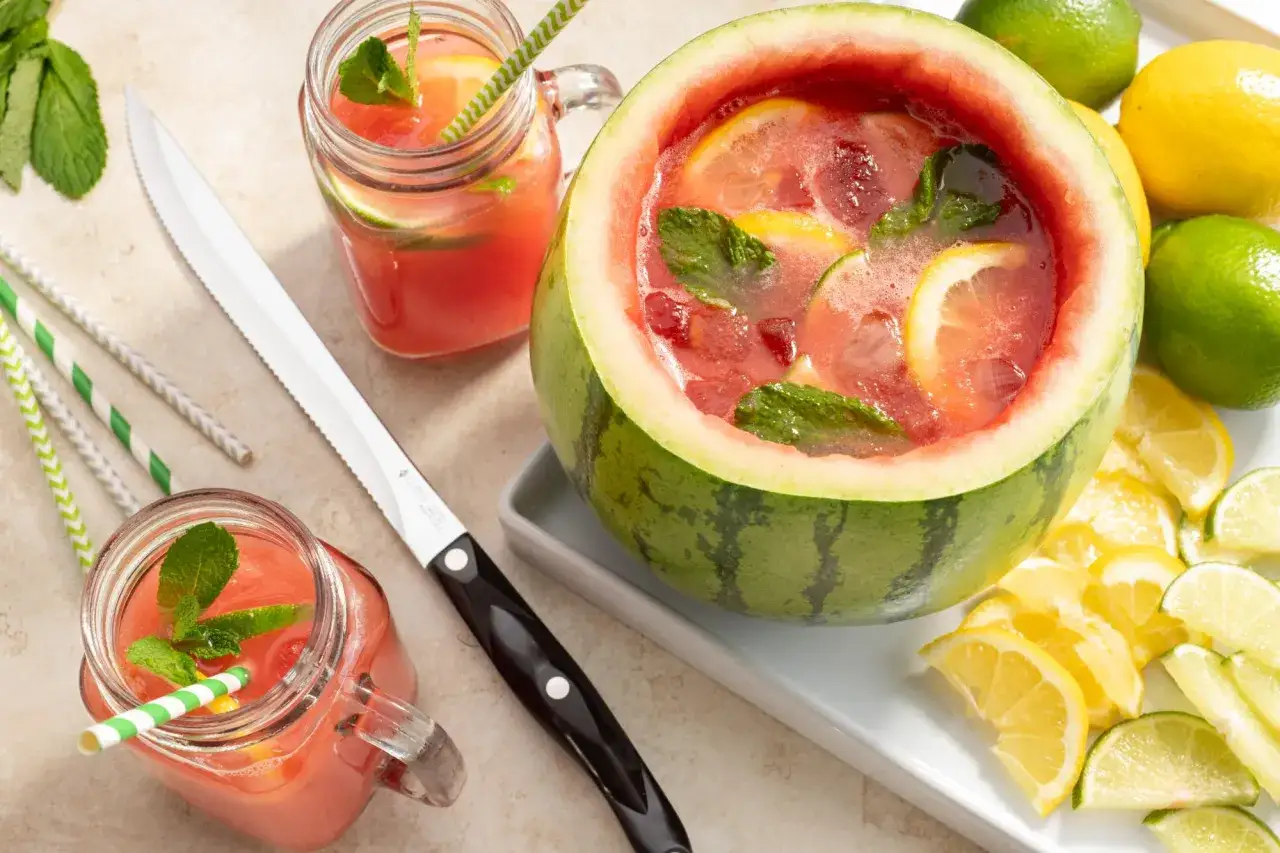 Orzeźwiająca lemoniada arbuzowa podana w wydrążonym arbuzie, udekorowana miętą i plasterkami cytryny. Obok dwa słoiczki z napojem, cytryny, limonki i słomki.