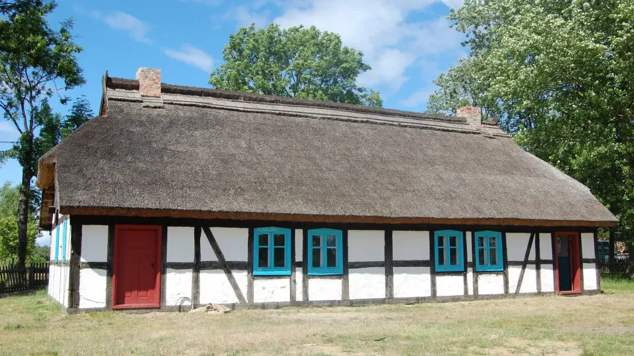 Skansen w Klukach: tradycyjny dom z bielonymi ścianami, czarną konstrukcją szachulcową i strzechą. Jasne okna i czerwone drzwi dodają uroku.