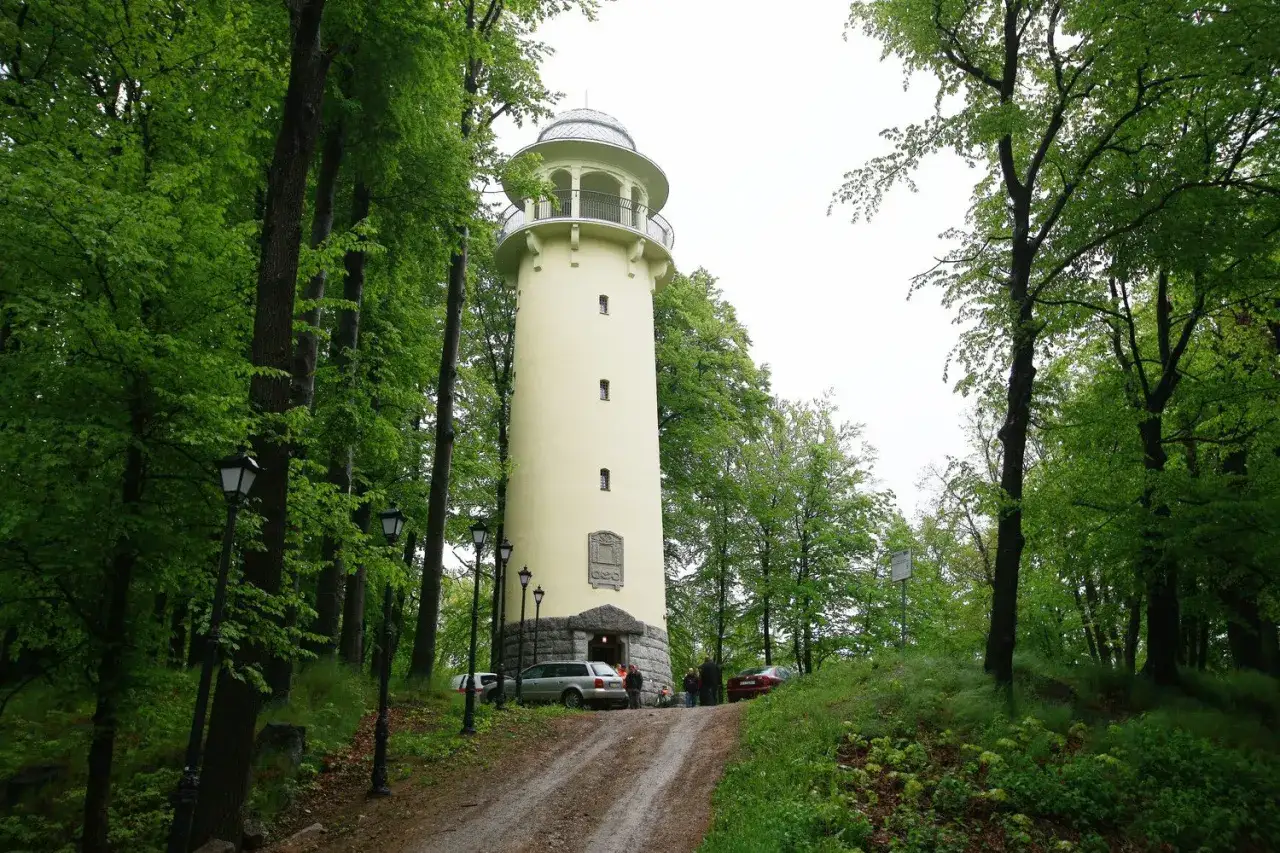 Jasnożółta wieża widokowa w Jeleniej Górze, otoczona bujną zielenią drzew. U podnóża wieży stoją samochody i kilka osób.
