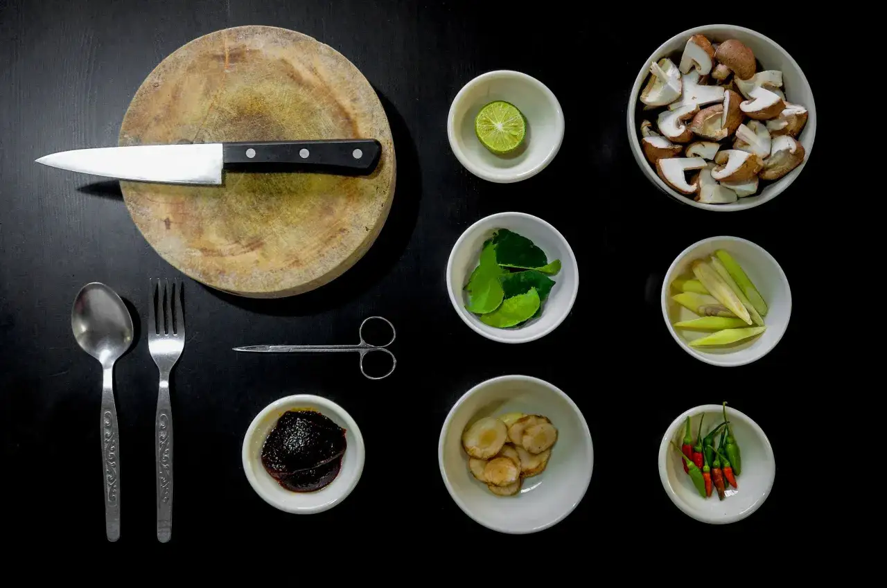 Mise and place en cocina: tabla, cuchillo, tenedor, cuchara, tijeras, champi&ntilde;ones, lima, hojas de lima kaffir, hierba lim&oacute;n, jengibre, pasta de chile y chiles.