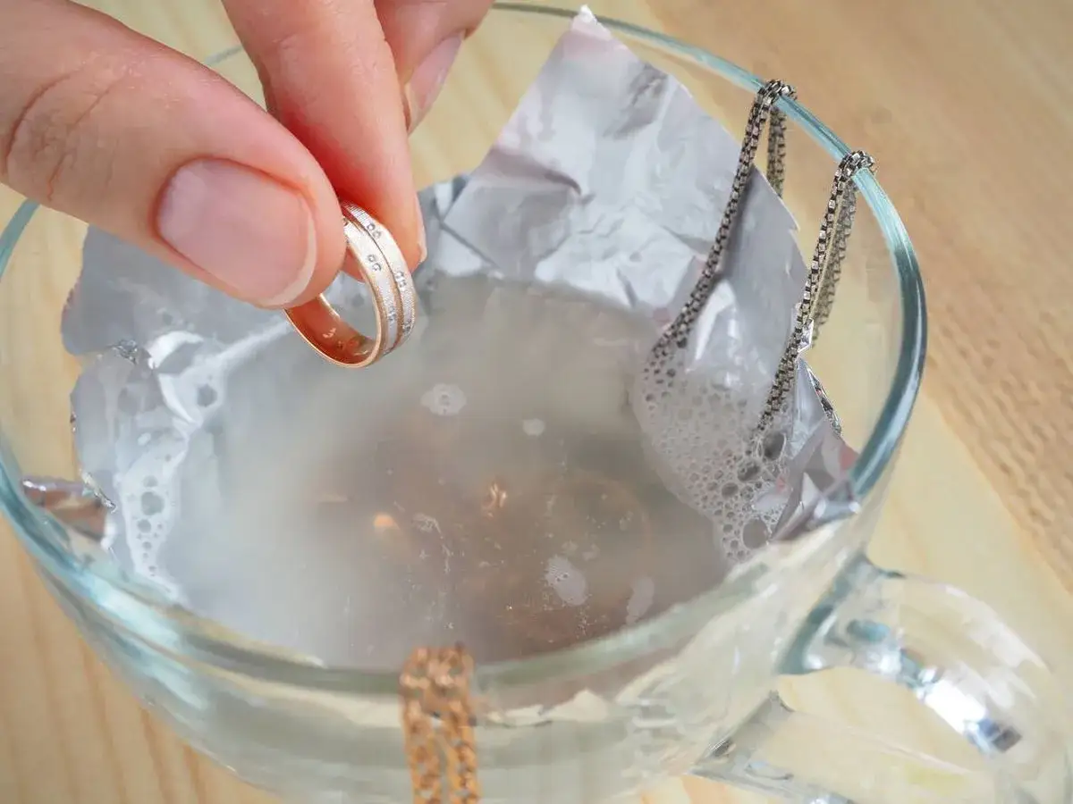 Mano sosteniendo un anillo de oro y plata, sumergi&eacute;ndolo en agua con burbujas para limpiar joyas. Una cadena plateada cuelga del borde del vaso.