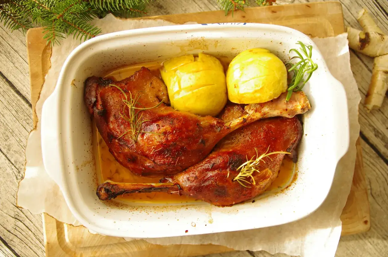 Pieczona gęsia nóżka z jabłkami i rozmarynem w białej brytfannie.