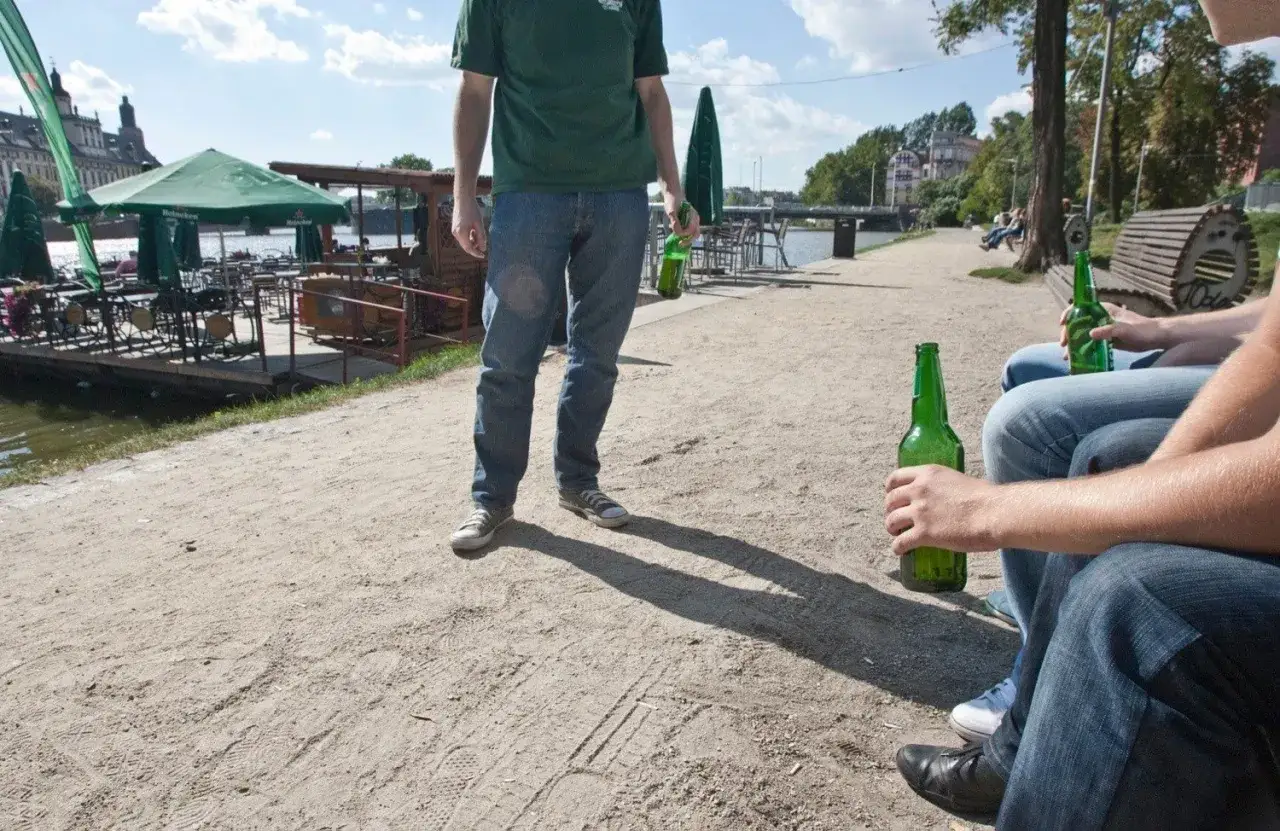 Ludzie piją piwo w miejscu publicznym nad rzeką, w tle widać restaurację na wodzie.