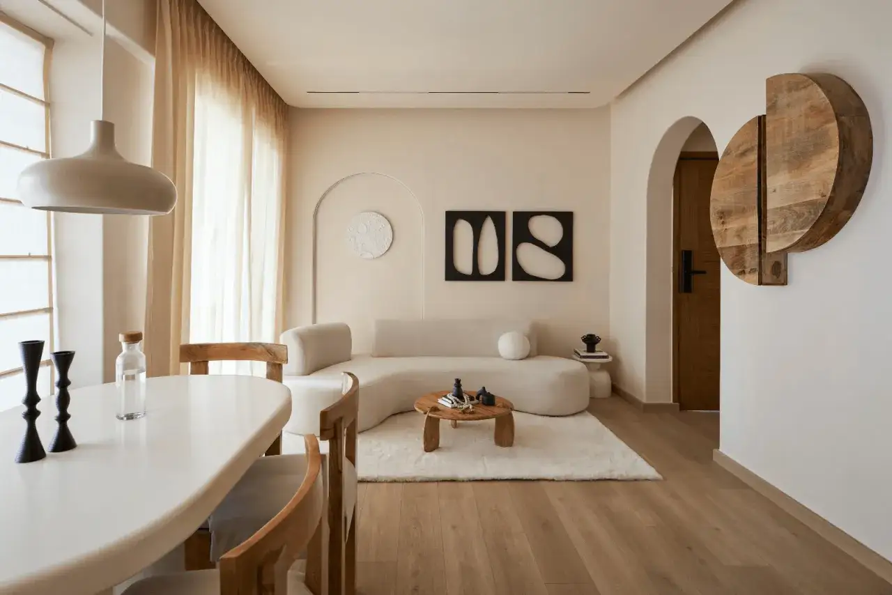 Ett vardagsrum med en mjuk, b&ouml;jd soffa och ett runt soffbord. Minimalistisk inredning med neutrala f&auml;rger och tr&auml;detaljer.