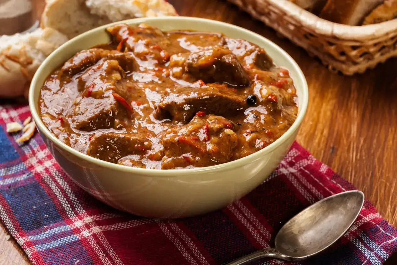 Aromaticzny gulasz z pręgi wołowej w misce, podany z chlebem i łyżką na kraciastej serwetce.