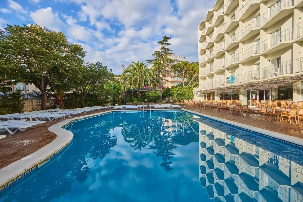Basen i taras z leżakami przy hotelu Don Juan Resort. Woda w basenie odbija chmury i budynek hotelu.