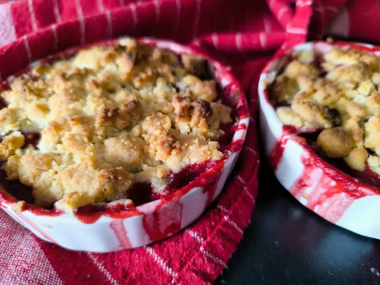 Deux ramequins de crumble fruits rouges, avec une croûte dorée et un cœur fruité, sur un tissu rouge à rayures.