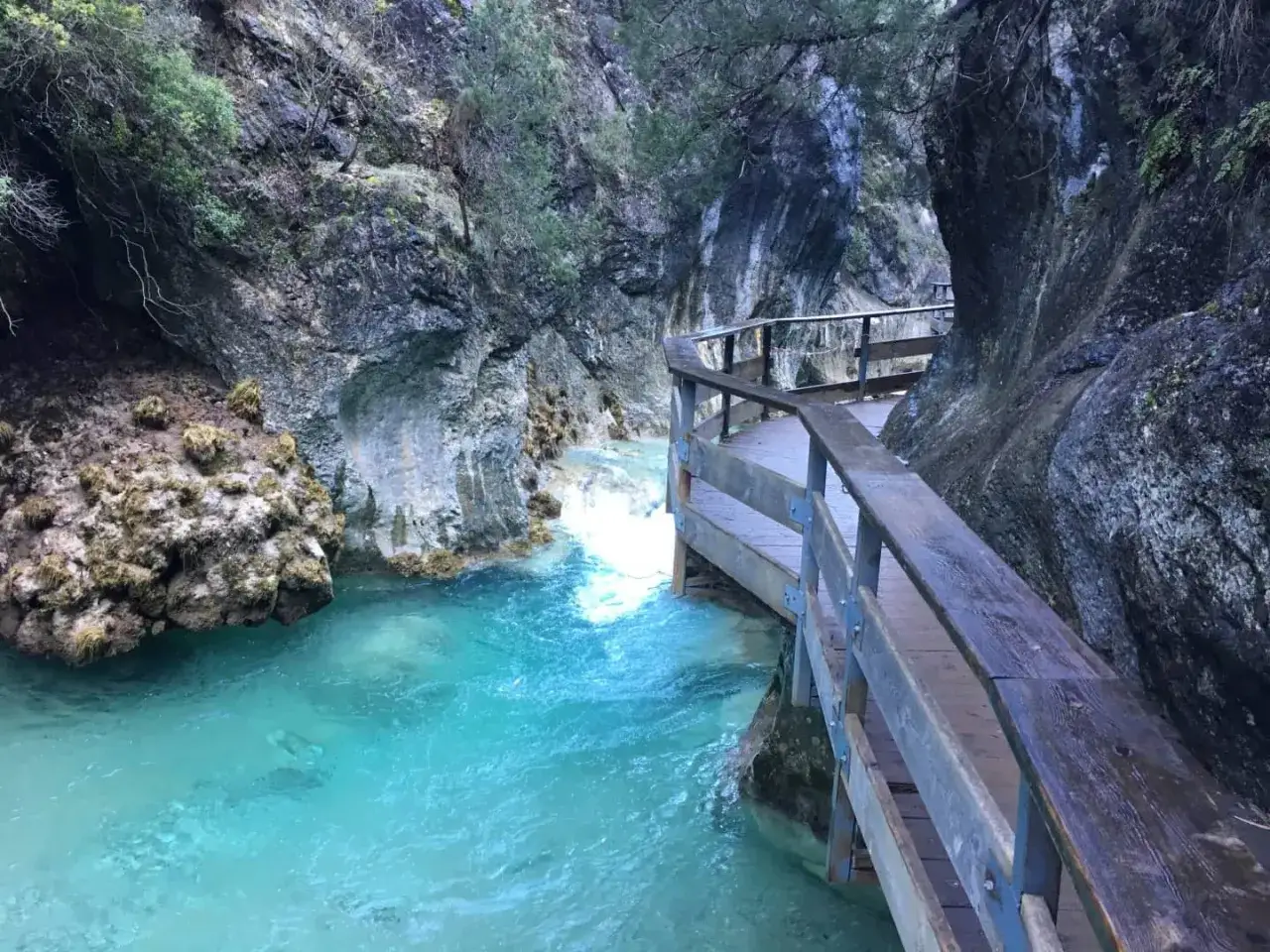 Pasarela de madera junto a un r&iacute;o turquesa en la **cerrada de El&iacute;as**. Rocas escarpadas y vegetaci&oacute;n cubren las paredes del ca&ntilde;&oacute;n.