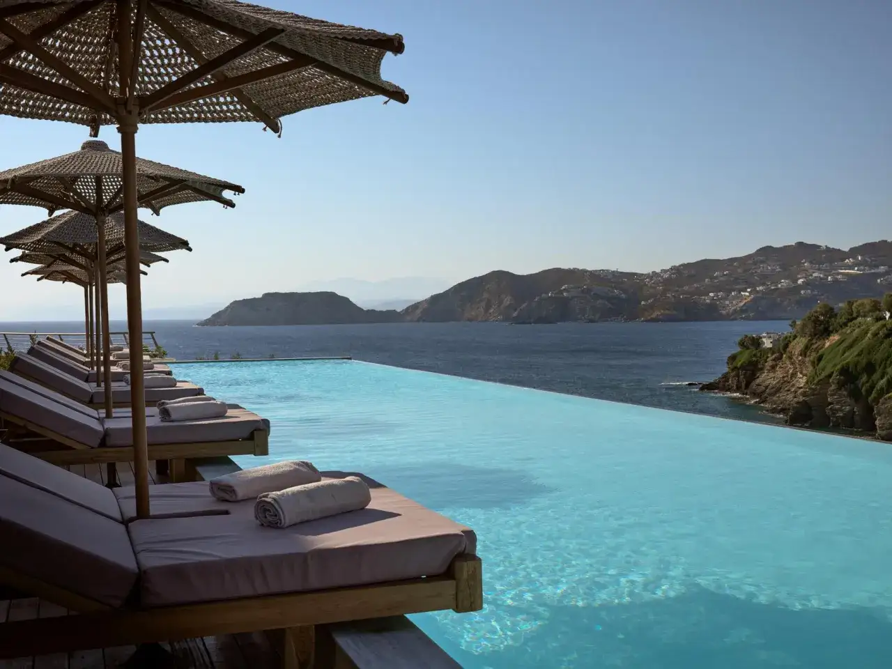 Entspannen Sie sich am Infinity-Pool des Arina Beach Resort. Liegestühle unter Sonnenschirmen laden zum Verweilen ein, während der Blick über das Meer zu den Hügeln schweift.