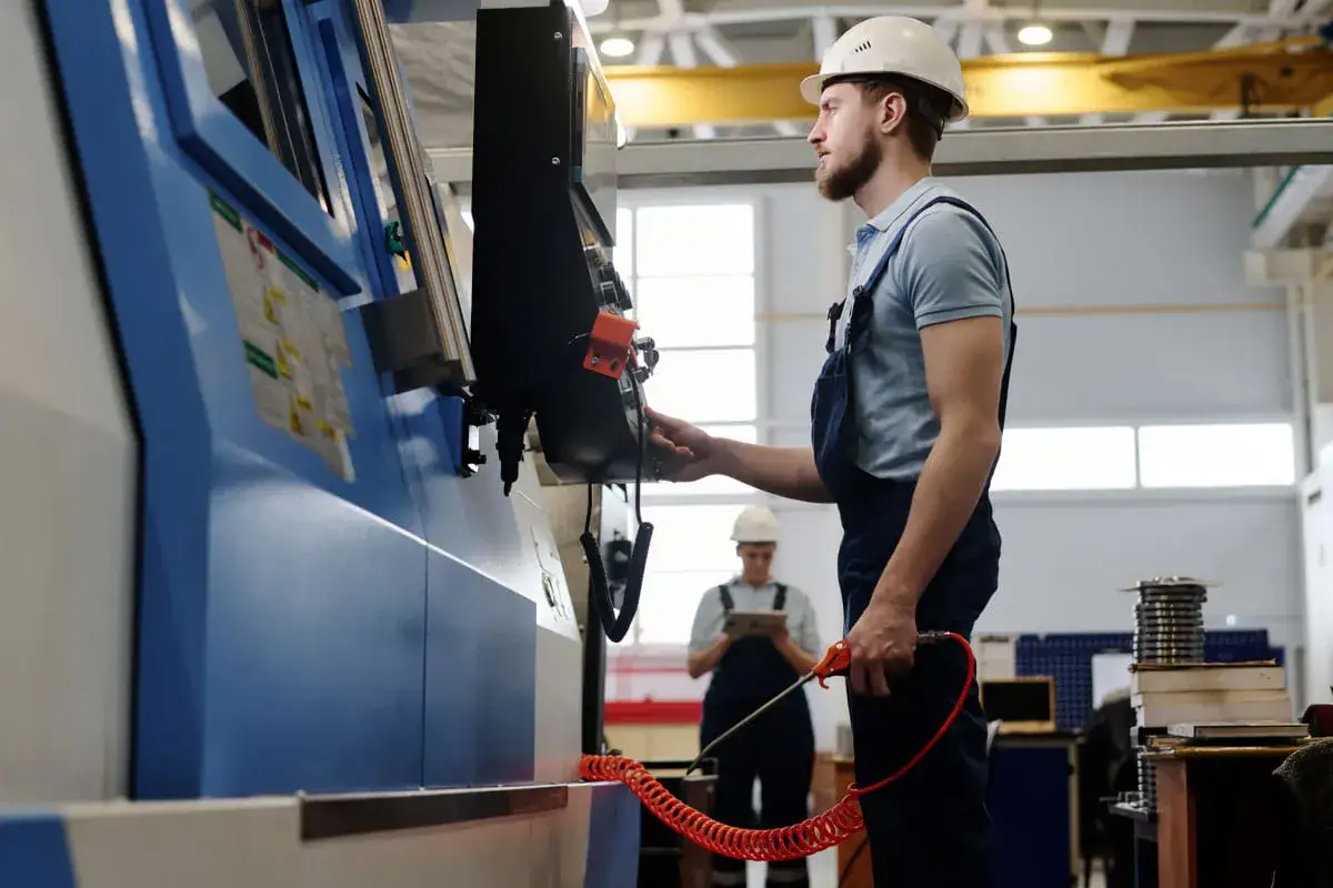 Operator maszyn CNC praca: mężczyzna w kasku obsługuje panel sterowania maszyny, w tle inny pracownik z tabletem.