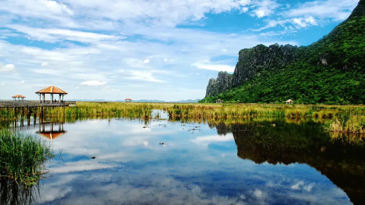 Khao Sam Roi Yot National Park: Ein ruhiger See spiegelt den Himmel und die üppige grüne Berglandschaft wider, mit kleinen Pavillons, die über dem Wasser liegen.