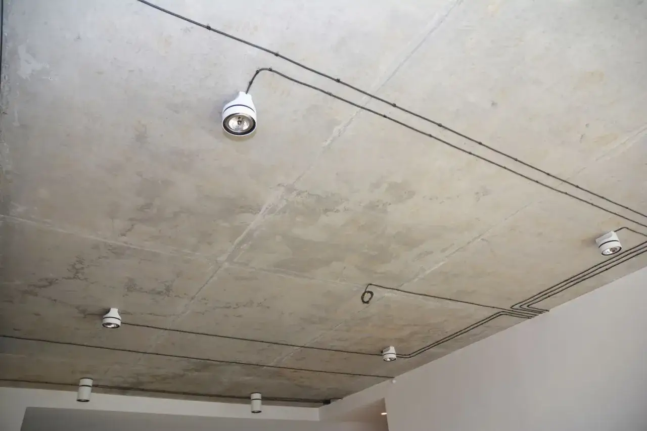 Nowoczesna wymiana instalacji elektrycznej bez kucia. Widoczne białe oprawy oświetleniowe i czarne przewody na betonowym suficie.