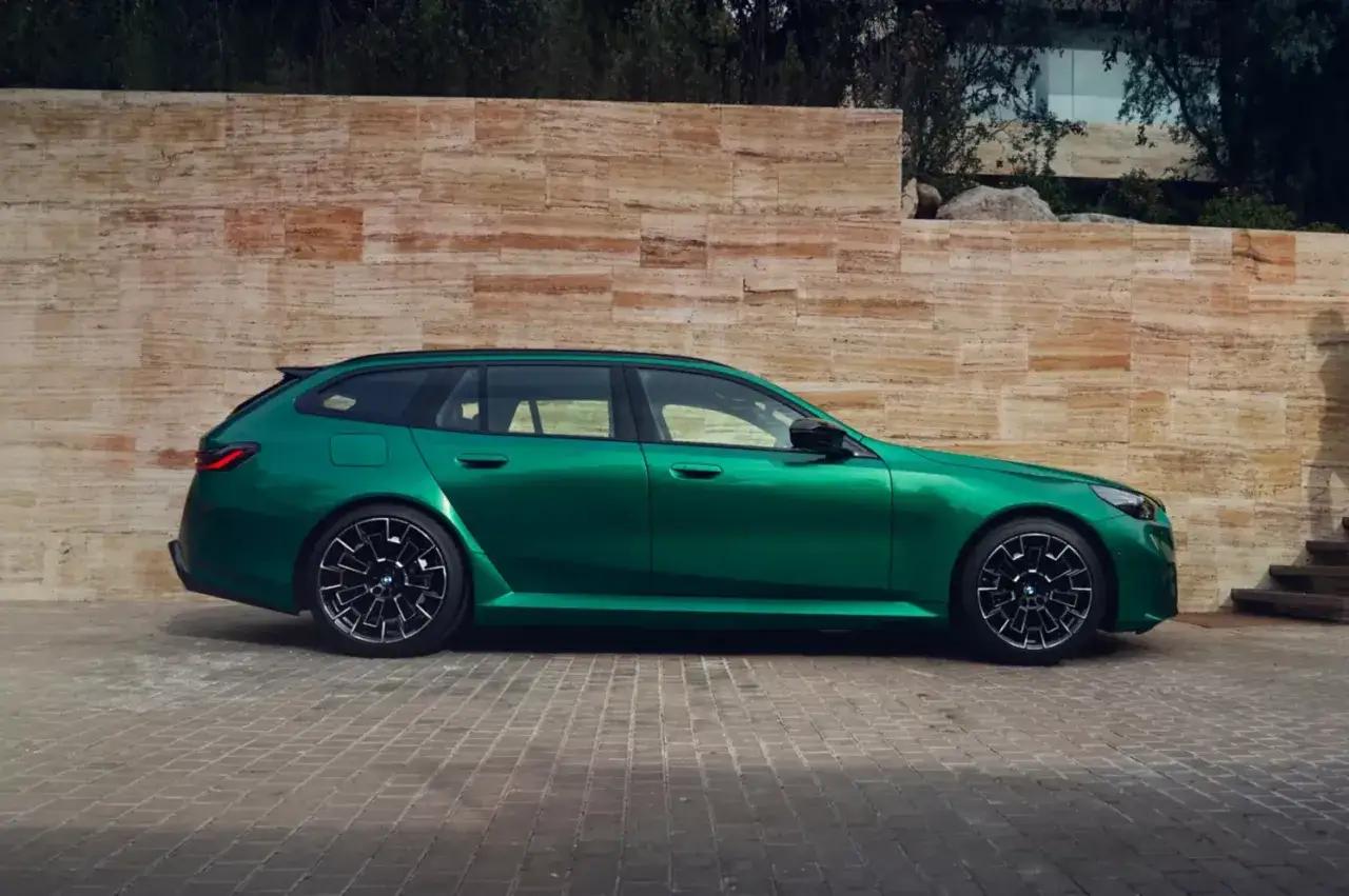 Zielony m5 touring lśni w słońcu, zaparkowany przed kamienną ścianą.