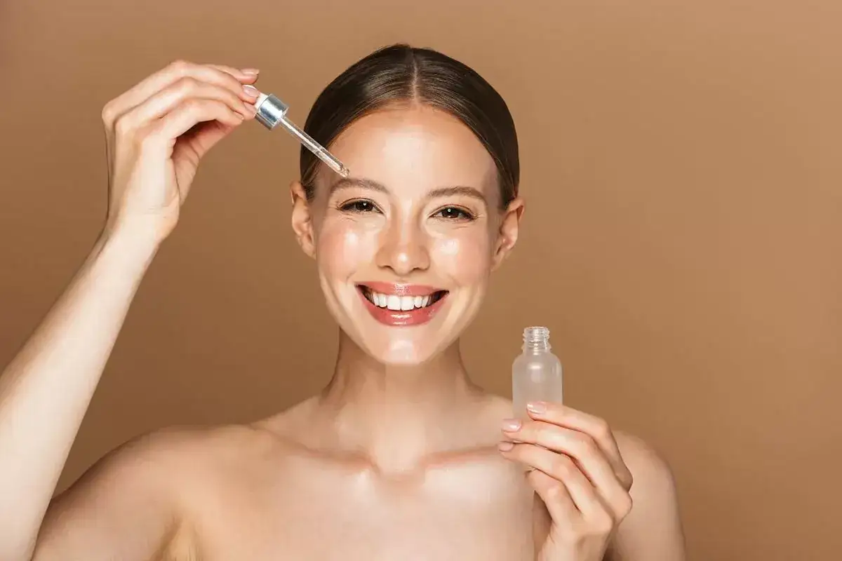 Una mujer sonr&iacute;e mientras se aplica serum en la frente con un gotero. Muestra c&oacute;mo se aplica el serum para una piel radiante.