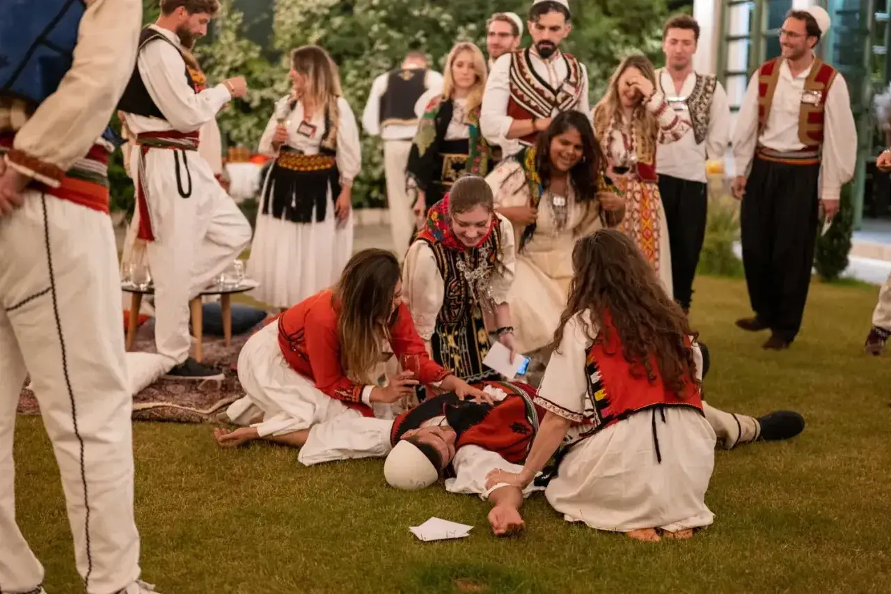 Ludzie w tradycyjnych strojach, być może podczas obchodzenia albańskich zwyczajów. Jedna osoba leży na ziemi, otoczona przez innych.