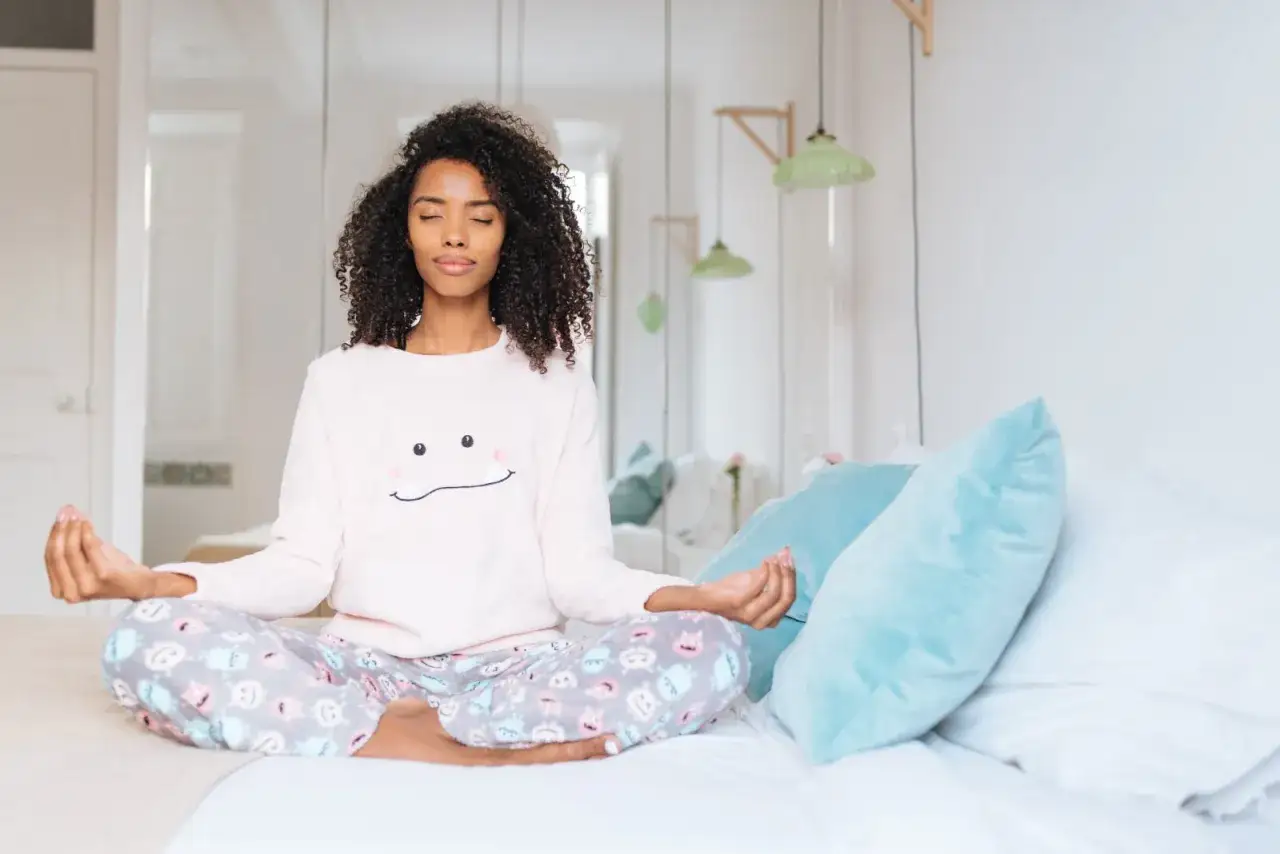 Eine Frau meditiert im Lotussitz auf einem Bett. Sie trägt einen Pyjama mit einem lächelnden Gesicht. Sie strahlt Ruhe aus, ein Bild von zufriedenen Menschen.