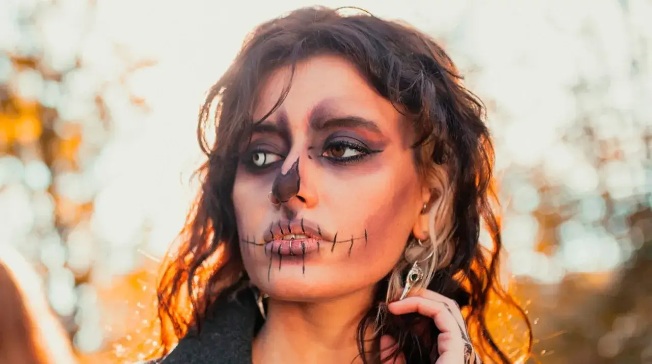 Makijaż na Halloween w stylu czaszki, z czarnymi szwami na ustach i oczach. Luźne, falowane włosy dopełniają przerażający wygląd.