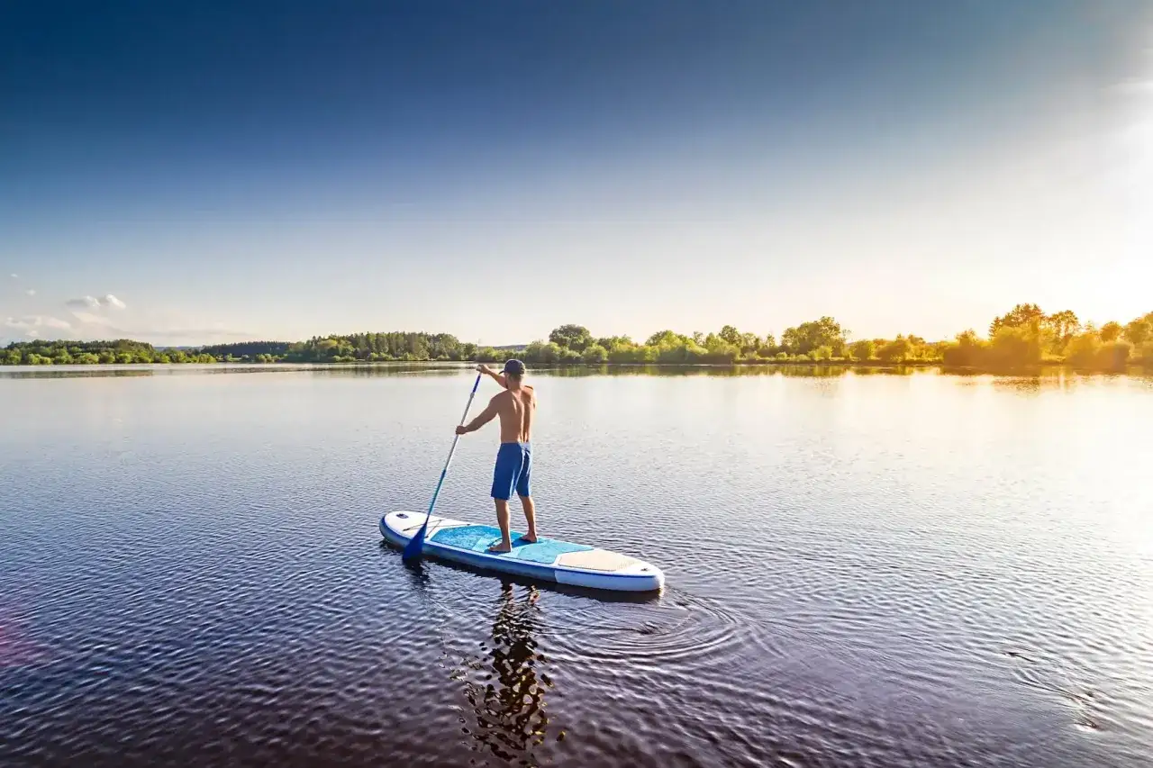 Mann auf SUP-Board auf ruhigem See, umgeben von gr&uuml;nen Ufern. Perfekt, um zu entspannen und die Natur zu genie&szlig;en. Hier darf man SUP fahren!