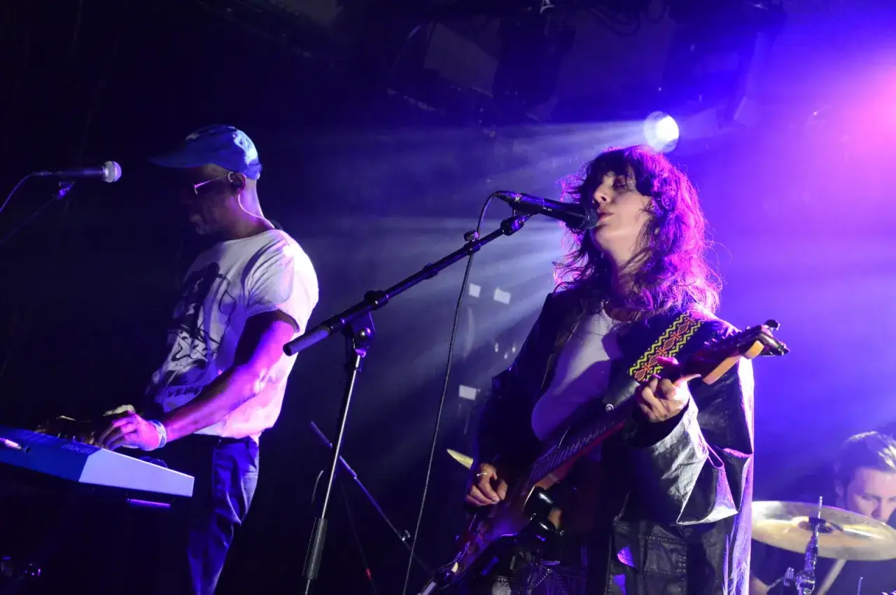 Une chanteuse joue de la guitare sous des lumières violettes, tandis qu'un homme à casquette bleue joue du clavier, créant une ambiance de concert dj house.