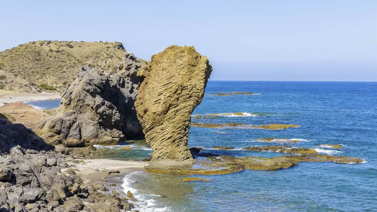 Roca imponente en una sopalmo playa, con el mar azul y rocas emergiendo del agua.