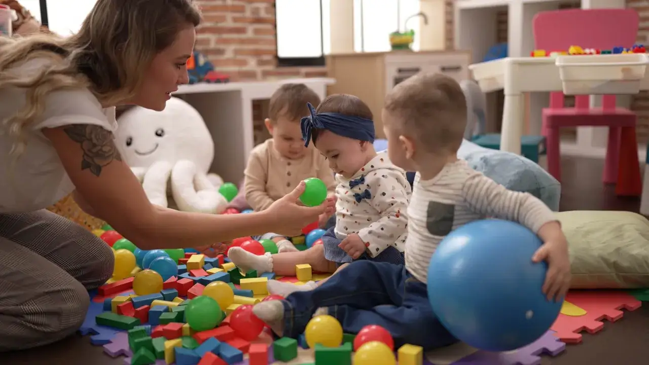 Nauczycielka bawi się z dziećmi klockami i piłkami. Czy żłobek państwowy jest płatny? Dzieci uczą się i bawią w przyjaznym otoczeniu.
