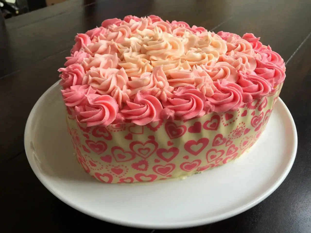 G&acirc;teau en forme de c&oelig;ur d&eacute;cor&eacute; de cr&egrave;me au beurre facile rose et p&ecirc;che, entour&eacute; d'une bande de chocolat blanc orn&eacute;e de c&oelig;urs roses.