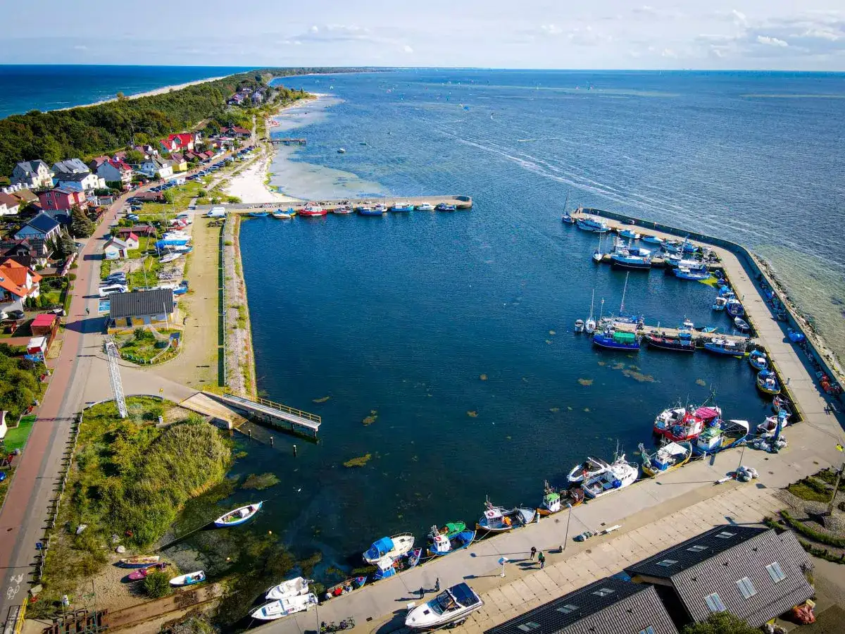 Widok z lotu ptaka na malowniczą **kuźnicę** z portem pełnym łodzi, domkami i piaszczystą plażą nad Bałtykiem.