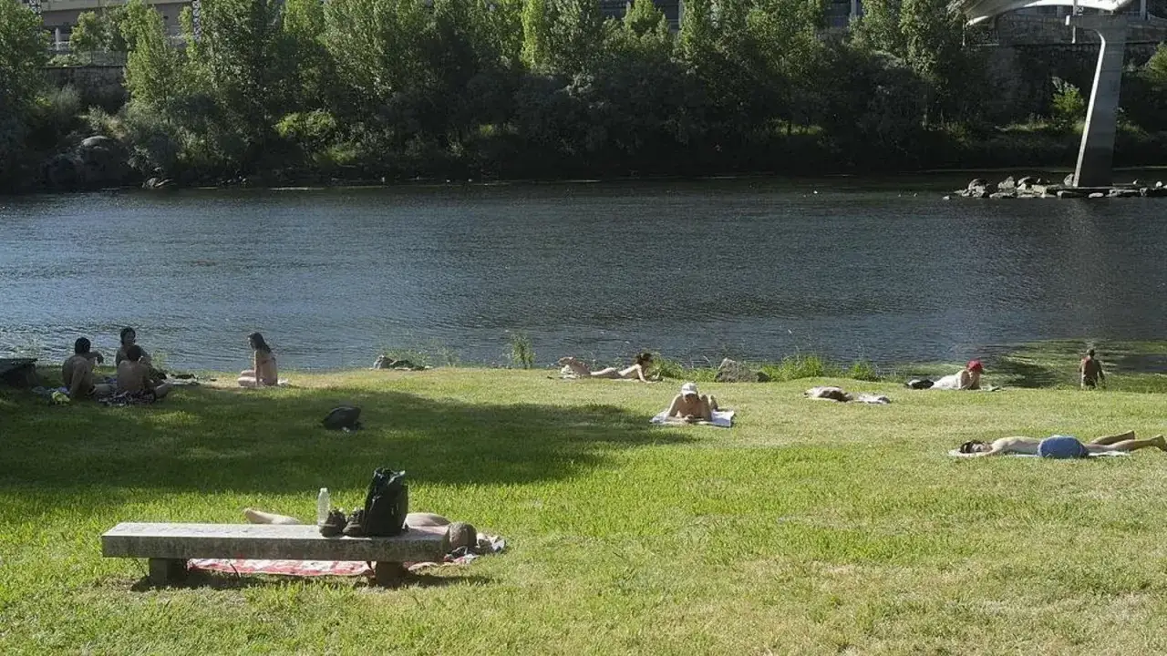Gente disfrutando del sol en la playa fluvial de Ourense, con el r&iacute;o Mi&ntilde;o de fondo y &aacute;rboles frondosos.
