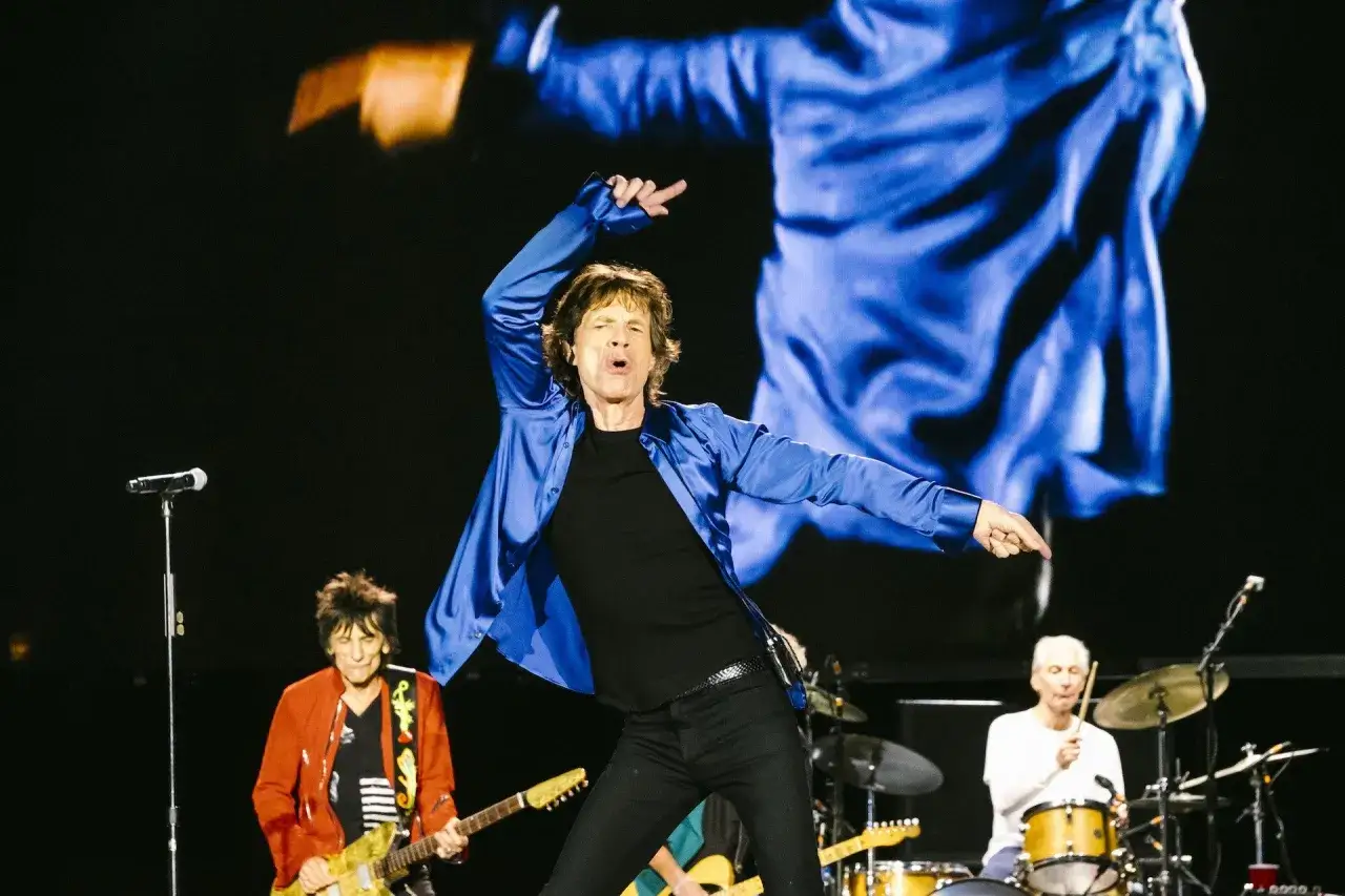 Mick Jagger w niebieskiej koszuli tańczy na scenie podczas jednego z koncert&oacute;w The Rolling Stones. Obok Ronnie Wood gra na gitarze, a Charlie Watts przy perkusji.