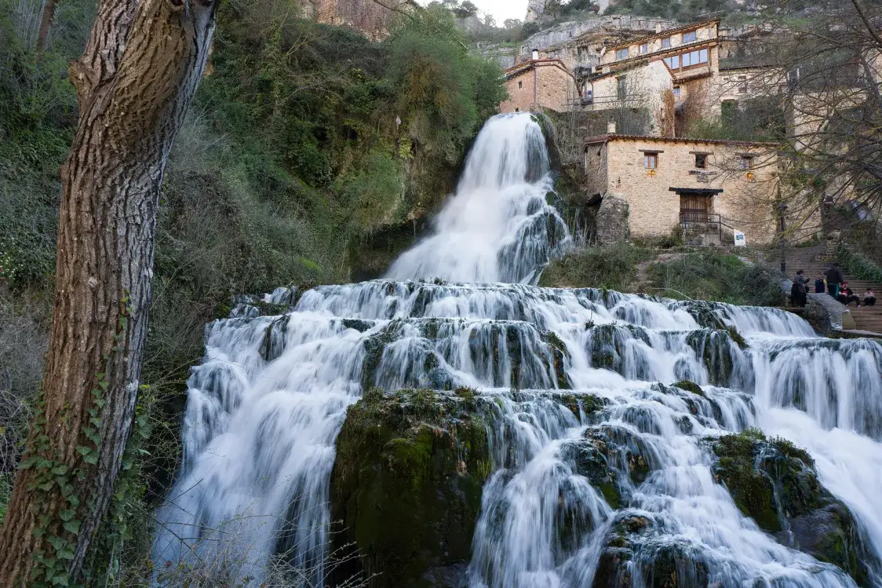Tosender Wasserfall st&uuml;rzt &uuml;ber Felsen, w&auml;hrend sich im Hintergrund die malerische Orbaneja del Castillo mit ihren Steinh&auml;usern an den Hang schmiegt.