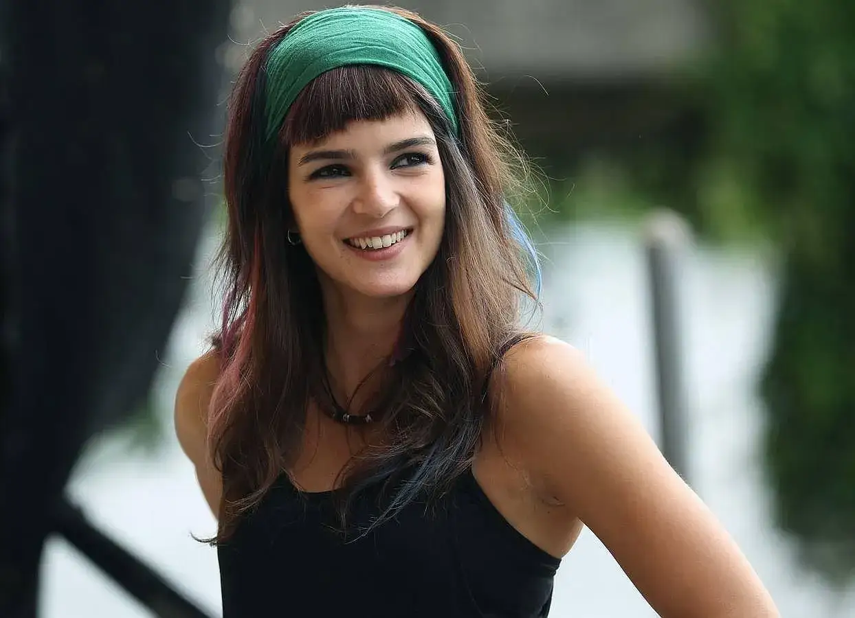 Mujer sonriente con un pañuelo verde y un flequillo vasco, con mechones de colores en su cabello castaño.